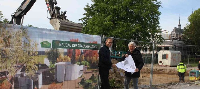 Architekt Peter Nikelowski vom Gebäudemanagement und Dietmar Maaßen von der Friedhofsverwaltung stehen vor der Baustelle neben einem Banner mit einem Bild des neuen Tauerhauses