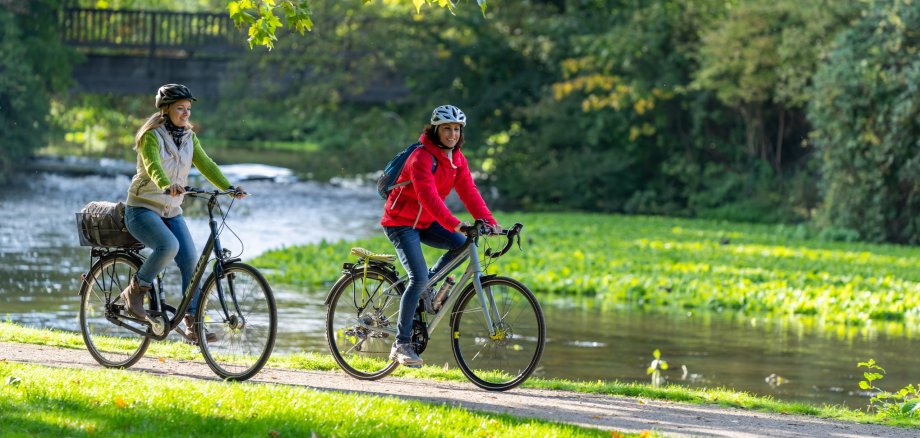 Zwei Personen fahren auf ihren Fahrrädern an einem Fluss entlang.